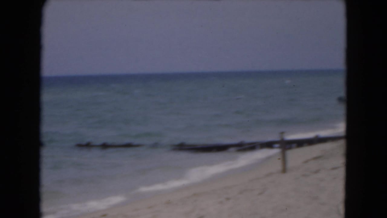 1975 Florida beach view with soft ocean surf, sand, wooden posts, and black 8mm projection borders.