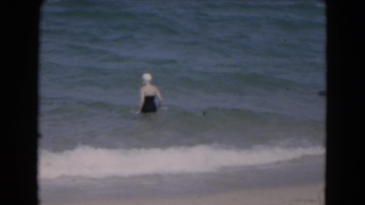 Woman in a dark swimsuit and white swim cap wading in shallow ocean water at a 1975 Florida beach.