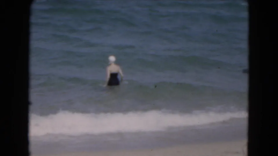 Woman in a dark swimsuit and white swim cap wading in shallow ocean water at a 1975 Florida beach.