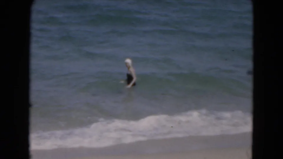A woman in a dark swimsuit and white cap stands in shallow Florida surf, filmed on soft 8mm home-movie footage.
