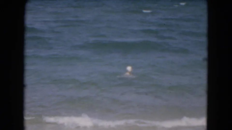 1975 Florida 8mm frame showing a distant swimmer in shallow ocean waves, viewed through dark vertical foreground framing.