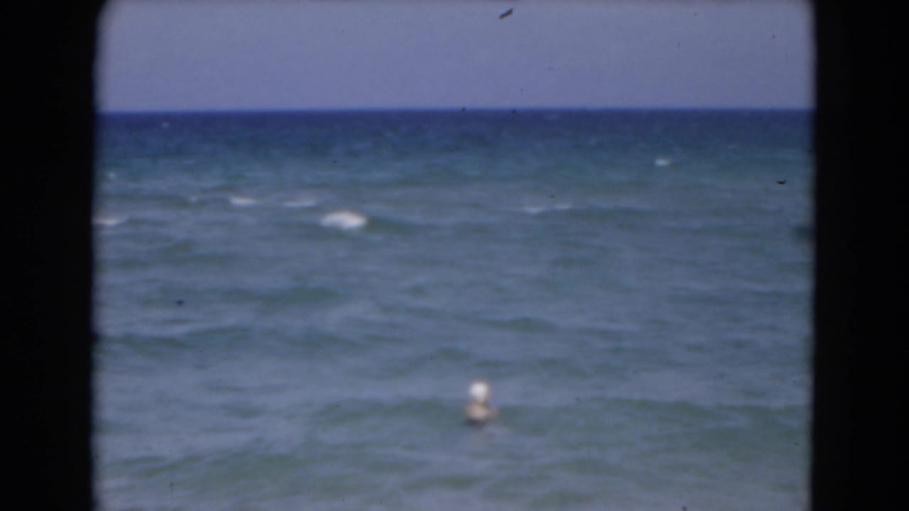 1975 Florida 8mm home-movie frame showing open ocean, low waves, pale sky, and a small white buoy offshore.