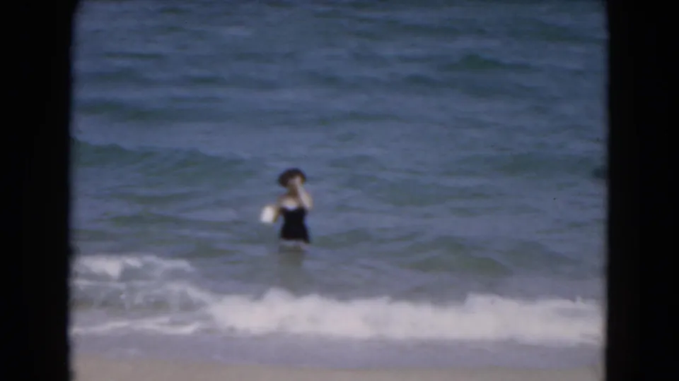 1975 Florida 8mm home-movie frame of a woman in a dark swimsuit wading in ocean surf near the beach.
