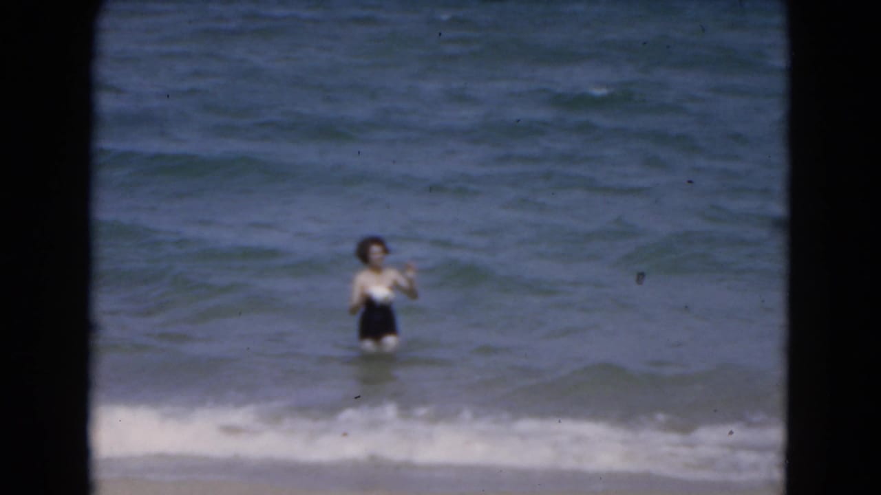 Woman in a black one-piece swimsuit standing in ocean surf near the beach.