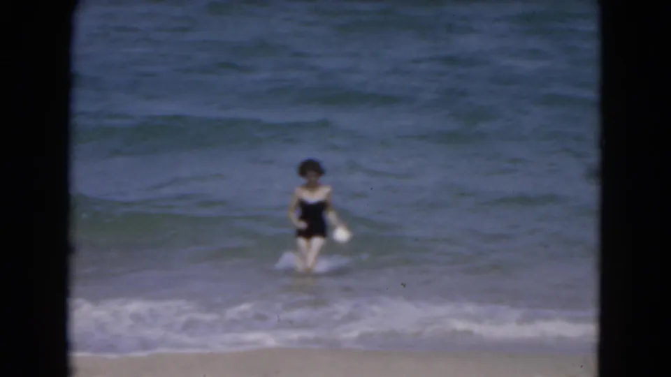 Adult woman walks through shallow Florida ocean waves, holding a white object, in 1975 8mm footage.