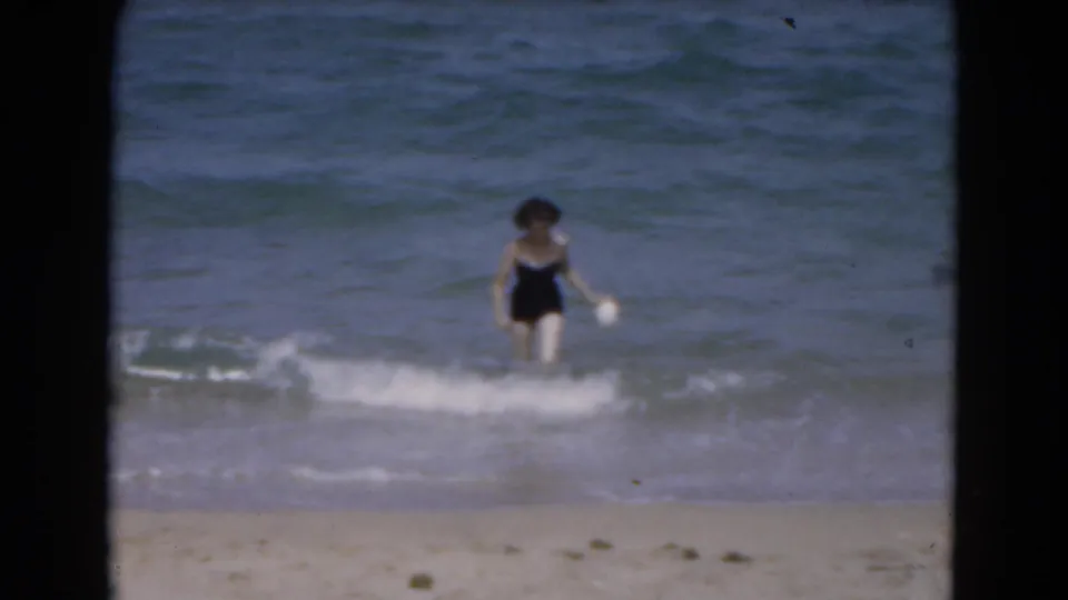 In 1975 Florida beach footage, an adult woman emerges from the ocean while white surf breaks around her legs.