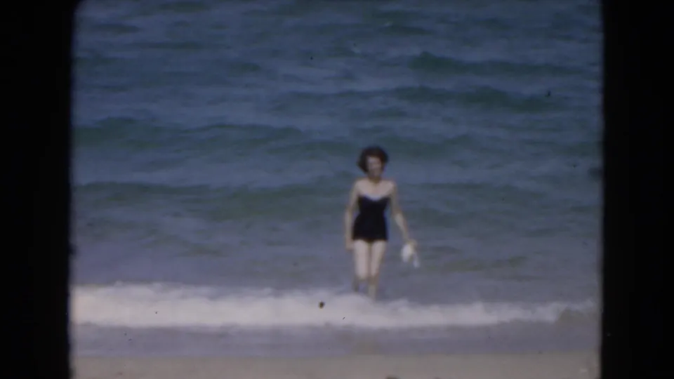 Adult woman in a black bathing suit walks toward the camera from Florida surf in 1975 home-movie footage.