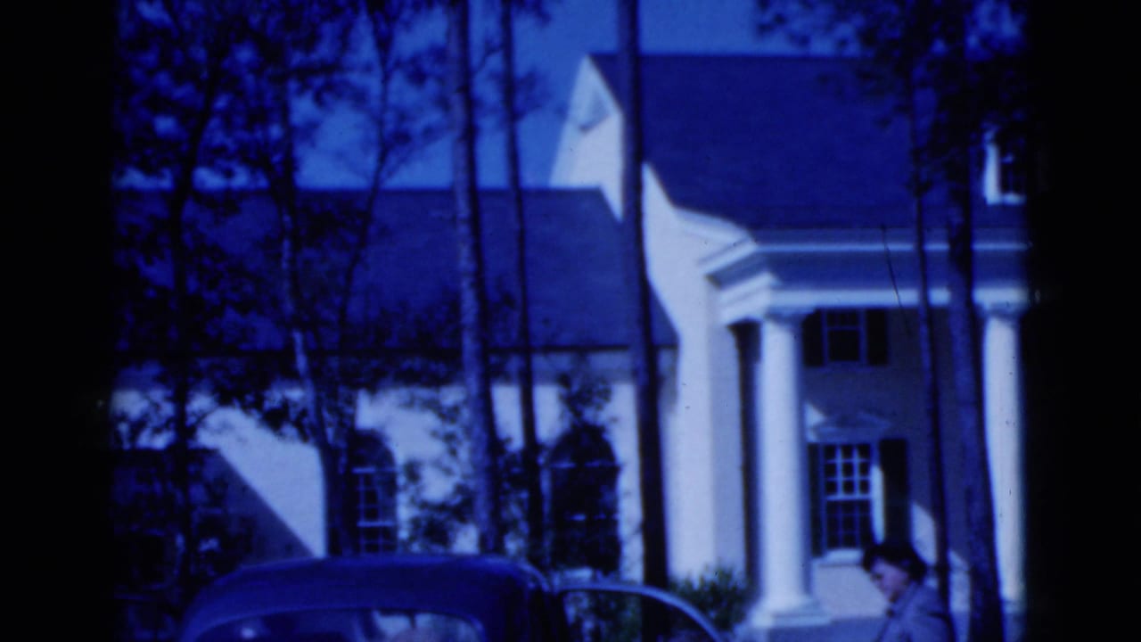 1975 Florida home-movie frame showing a white columned house partly obscured by trees, with a car parked in front.