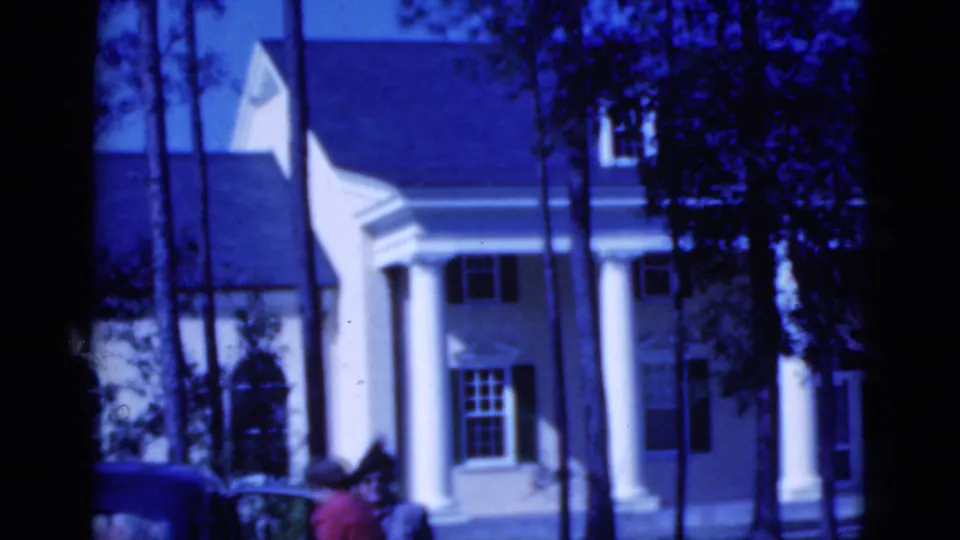 1975 Florida home-movie frame showing a woman in red near a parked car, with the white columned house behind her.