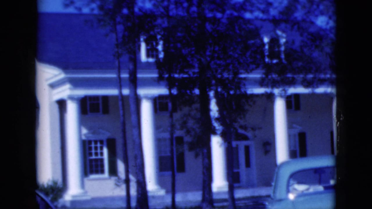 Columned house behind trees with a light-colored convertible partly visible in front.