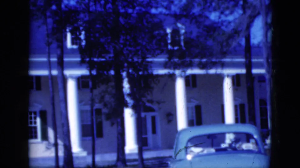 Blue-tinted Florida home-movie frame from 1975 with palm trunks, a columned house facade, and an older convertible in front.