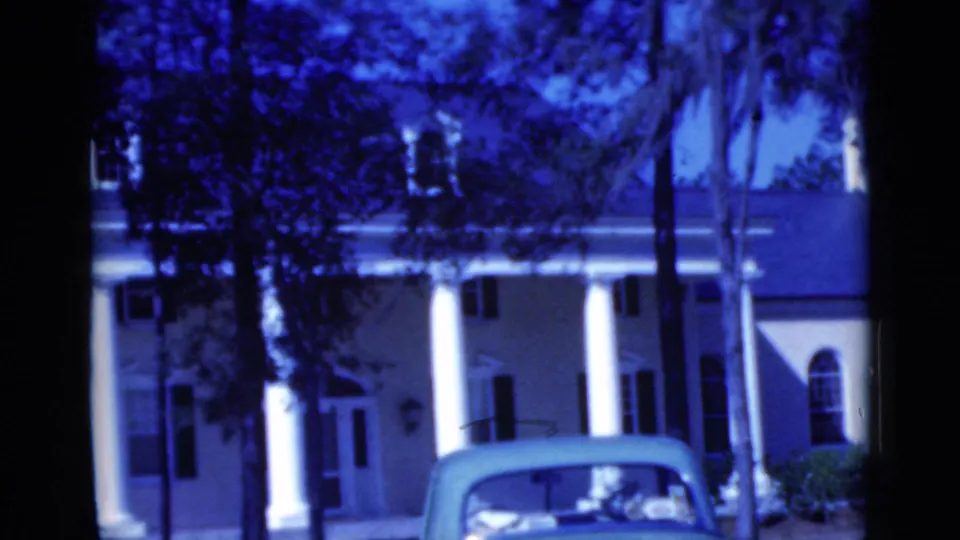 1975 Florida 8mm frame with a blue cast, showing a stately house, tall columns, palm trees, and a convertible in the foreground.