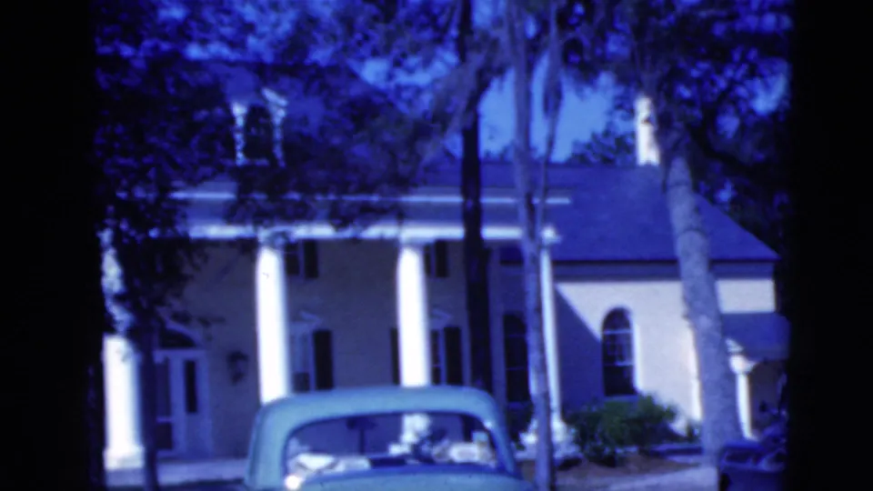 End frame of the 1975 Florida 8mm clip showing the columned house exterior, palm-lined yard, shrubs, and light-colored convertible.