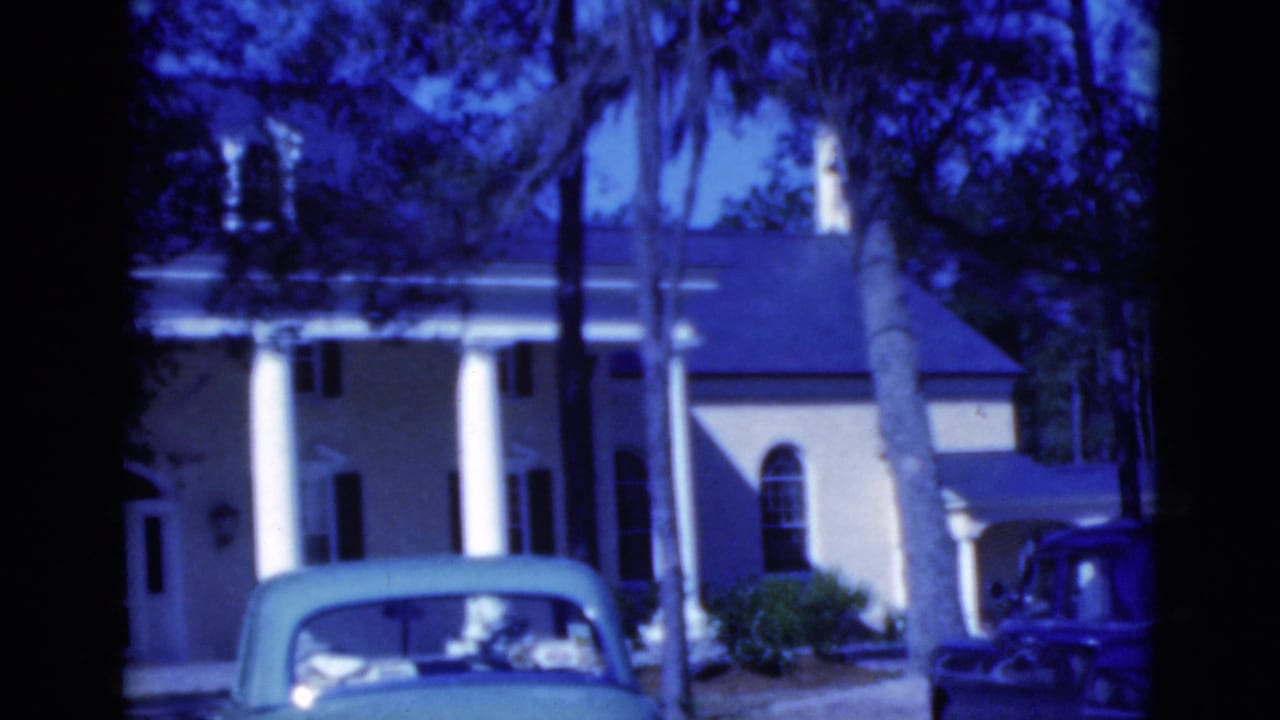 Columned house behind palms with a blue-green parked car in the foreground.