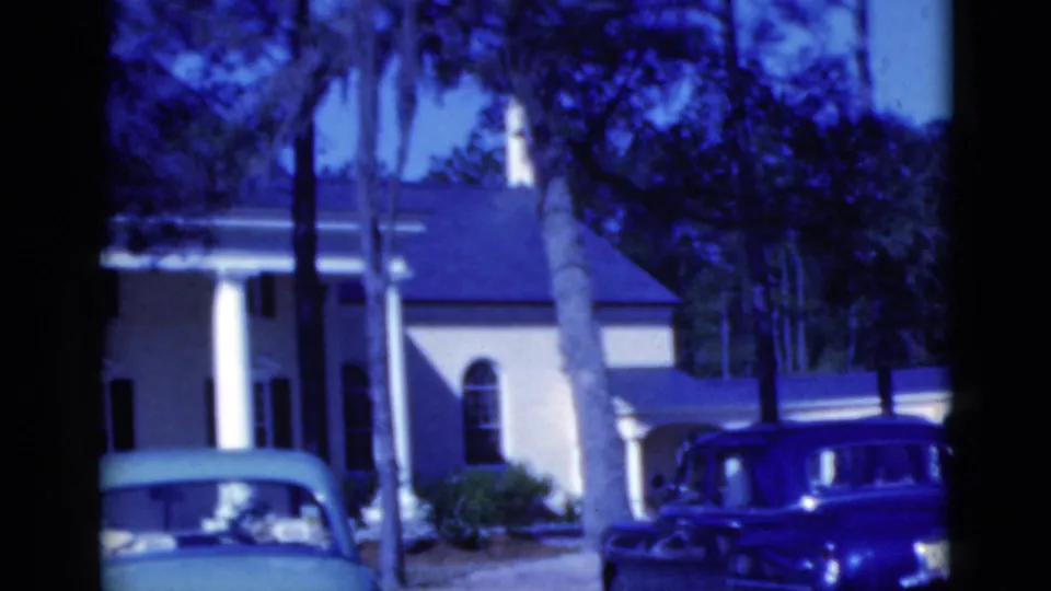1975 Florida home-movie frame with a columned house, tall palms, a blue-green car, and a dark station wagon in the driveway.