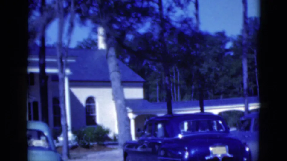 1975 Florida 8mm frame showing the side of a columned house, tall trees, shrubs, and a dark vintage station wagon.
