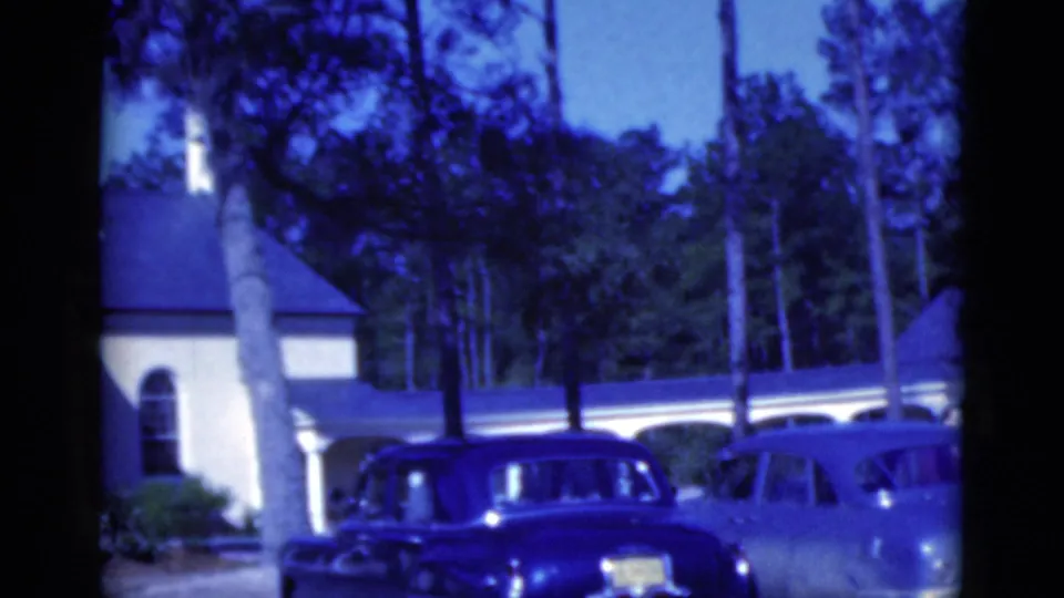 1975 Florida home-movie frame with a dark station wagon, pale car edge, columned house, palms, and dense trees.