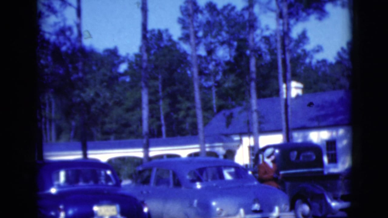 1975 Florida home-movie frame with sedans, a dark pickup truck, and a low white building in a pine-lined parking area.