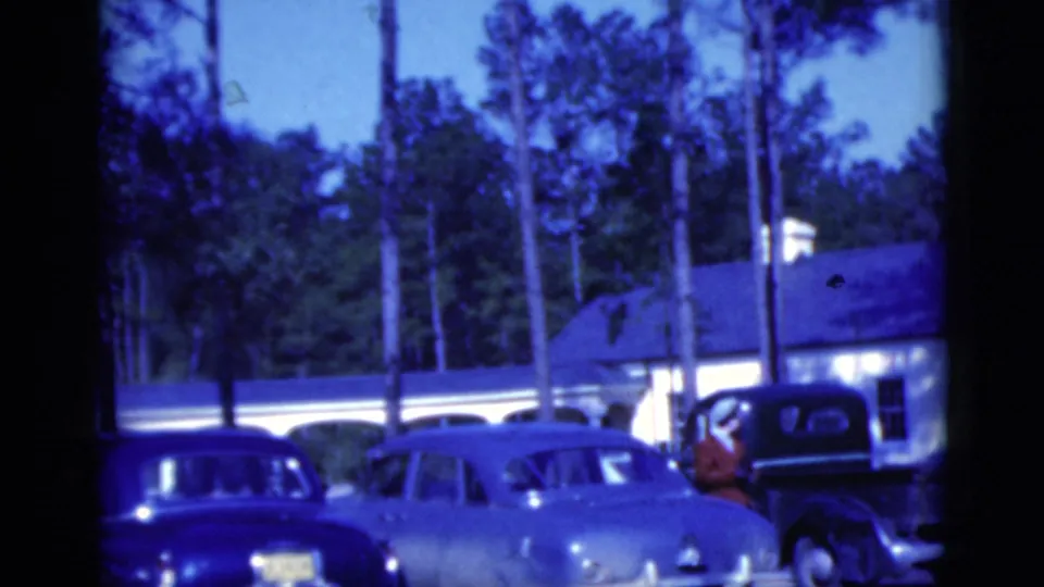1975 Florida home-movie frame with sedans, a dark pickup truck, and a low white building in a pine-lined parking area.