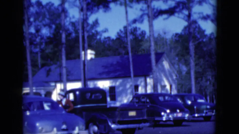 1975 Florida home-movie frame of a parking lot with several cars, a dark truck, pine trees, and a white building at dusk.