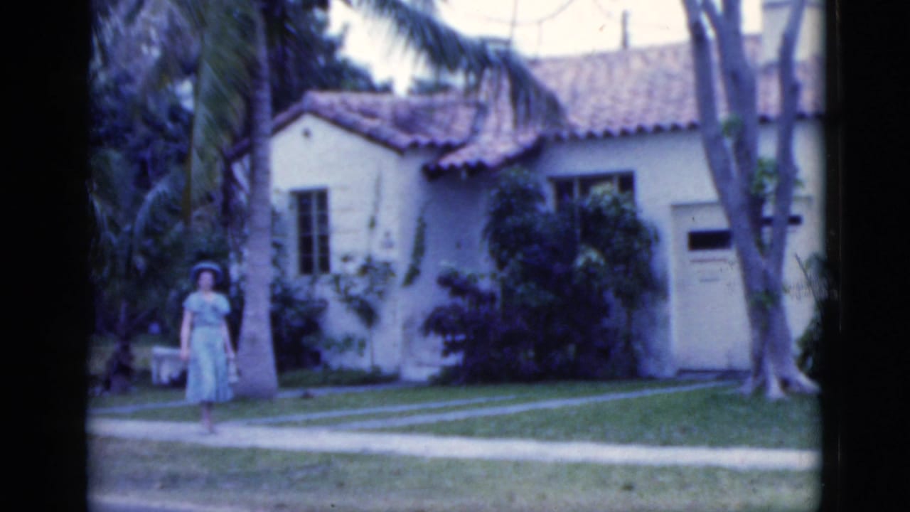 1975 Florida 8mm frame of a woman in a light blue sundress and hat approaching along a path outside a stucco home with palms.