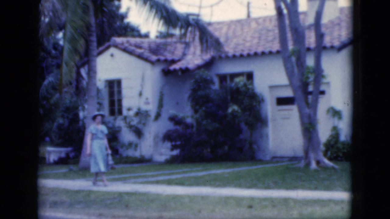 Woman in a hat walking beside a palm tree in front of a white stucco house.