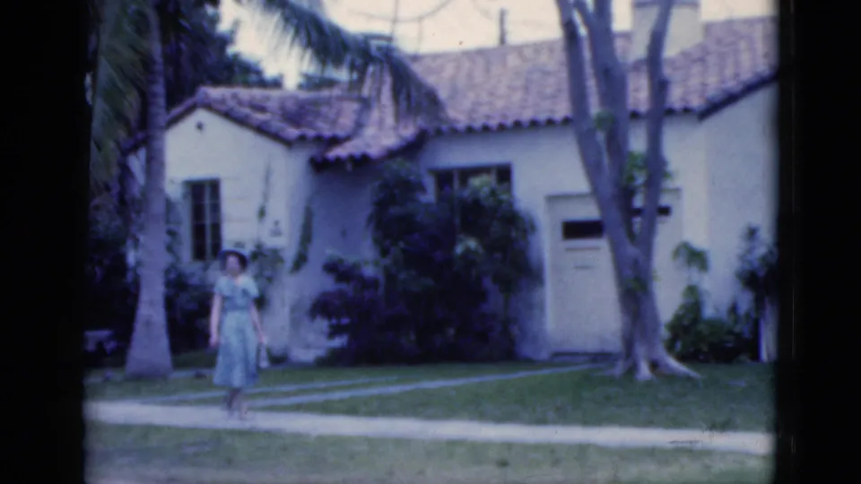 1975 Florida 8mm frame with a woman in a blue dress walking closer to camera near a palm tree and stucco residence.