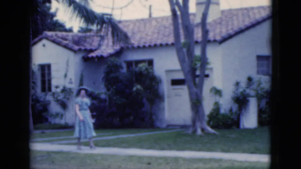 1975 Florida frame of a woman in a light blue sundress and hat near a sidewalk, with palms and a stucco home behind her.