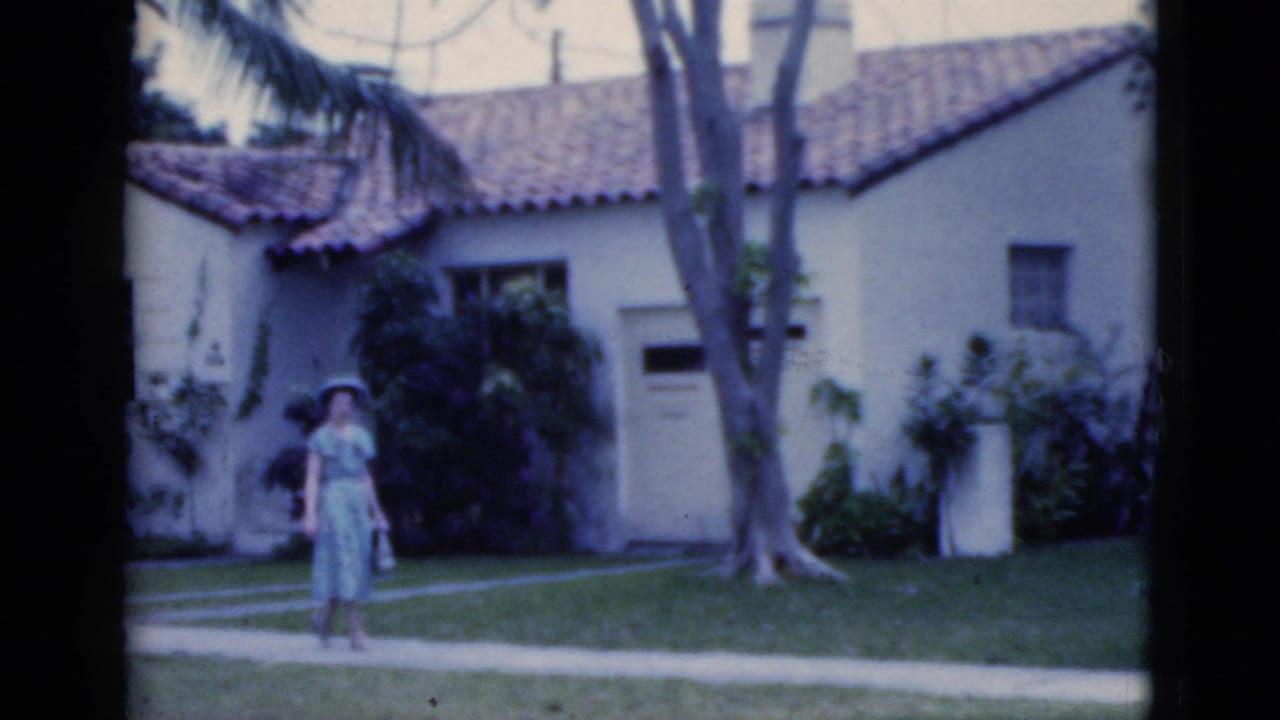 In 1975 Florida home-movie footage, a woman in a light blue dress and hat stands near a walkway in front of a Spanish-style house.