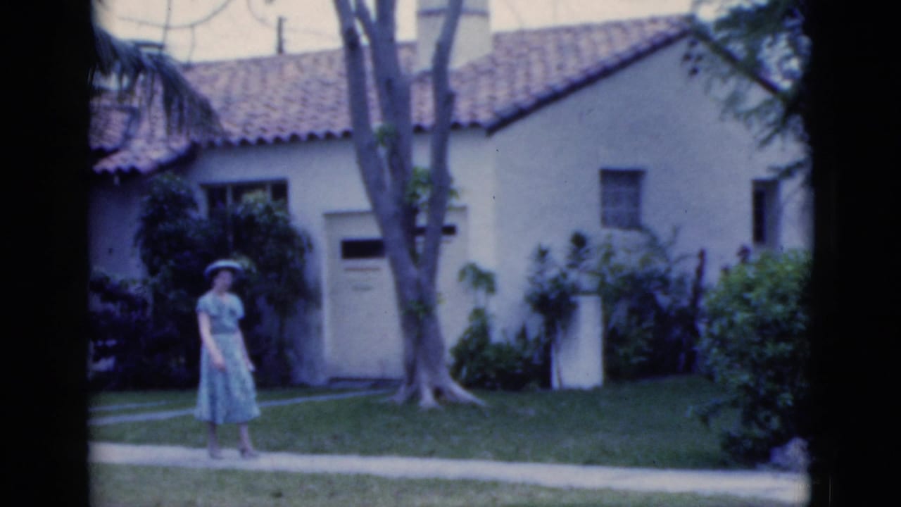Woman walks along a concrete path past a stucco house with trees and shrubs.