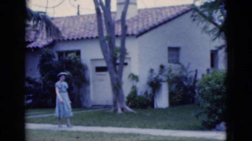 The woman moves along the walkway in front of the Spanish-style house, framed by lawn, shrubs, and tall tree trunks.
