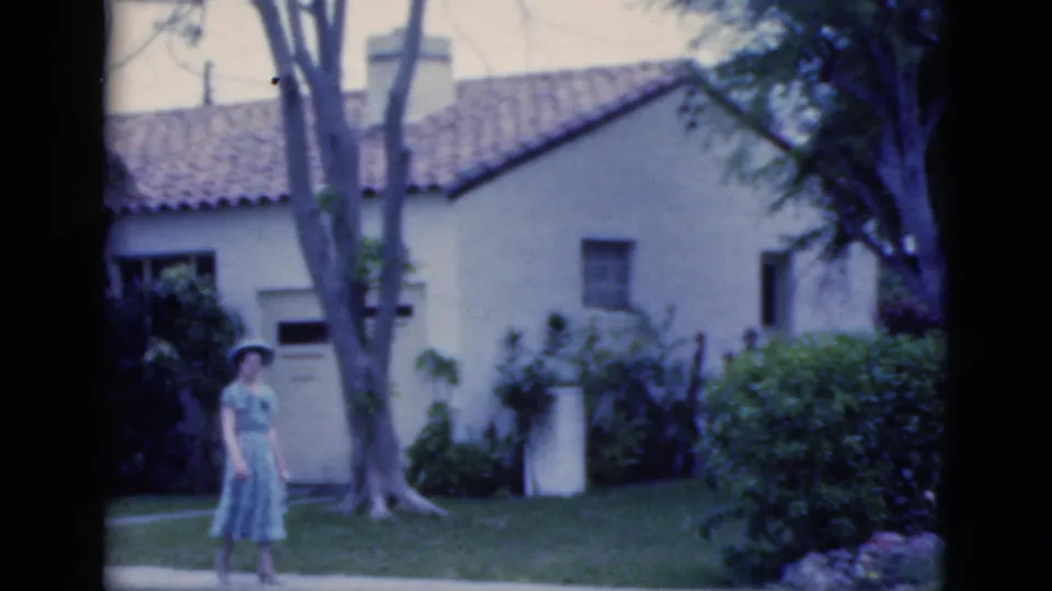 Mid-clip frame shows the woman continuing left to right along the path, with the stucco wall, roof tiles, lawn, and shrubs behind her.