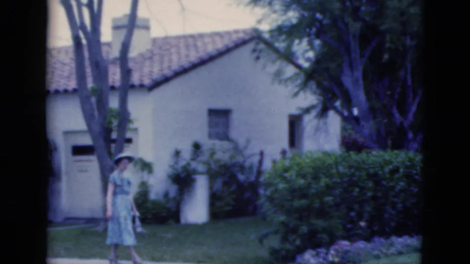 Near the end of the shot, the woman passes closer to the shrubs, with the Spanish-style house, lawn, and large trees still visible.