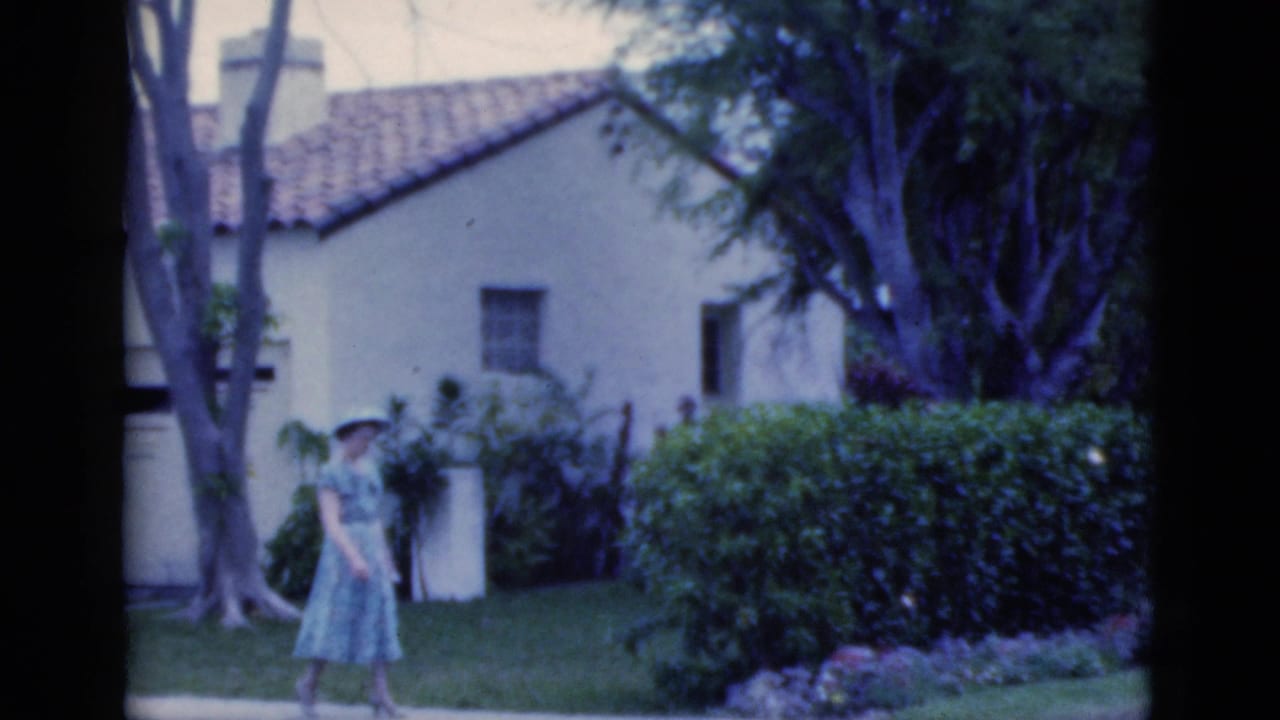 In 1975 Florida home-movie footage, a woman in a light blue dress and wide-brimmed hat walks past garden shrubs near a stucco house.