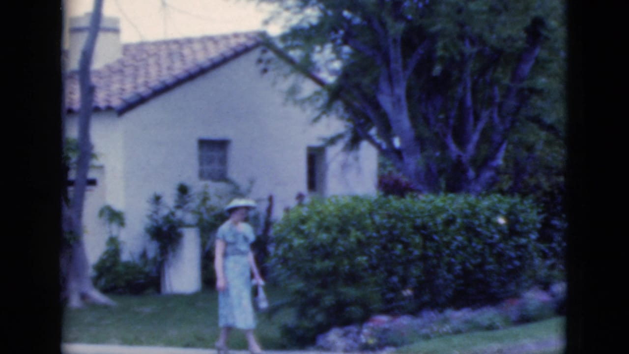 A woman in a light dress and hat continues along a garden path in front of a stucco house, consistent with the Florida 1975 clip metadata.