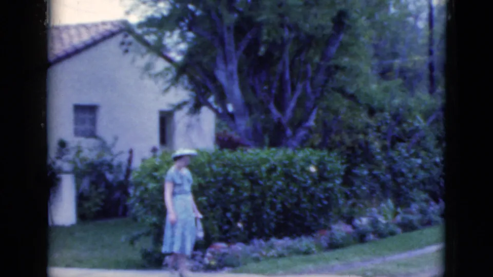 The woman in a light dress and hat moves beside a hedge and lawn, with a stucco house and mature trees visible in the 1975 Florida scene.