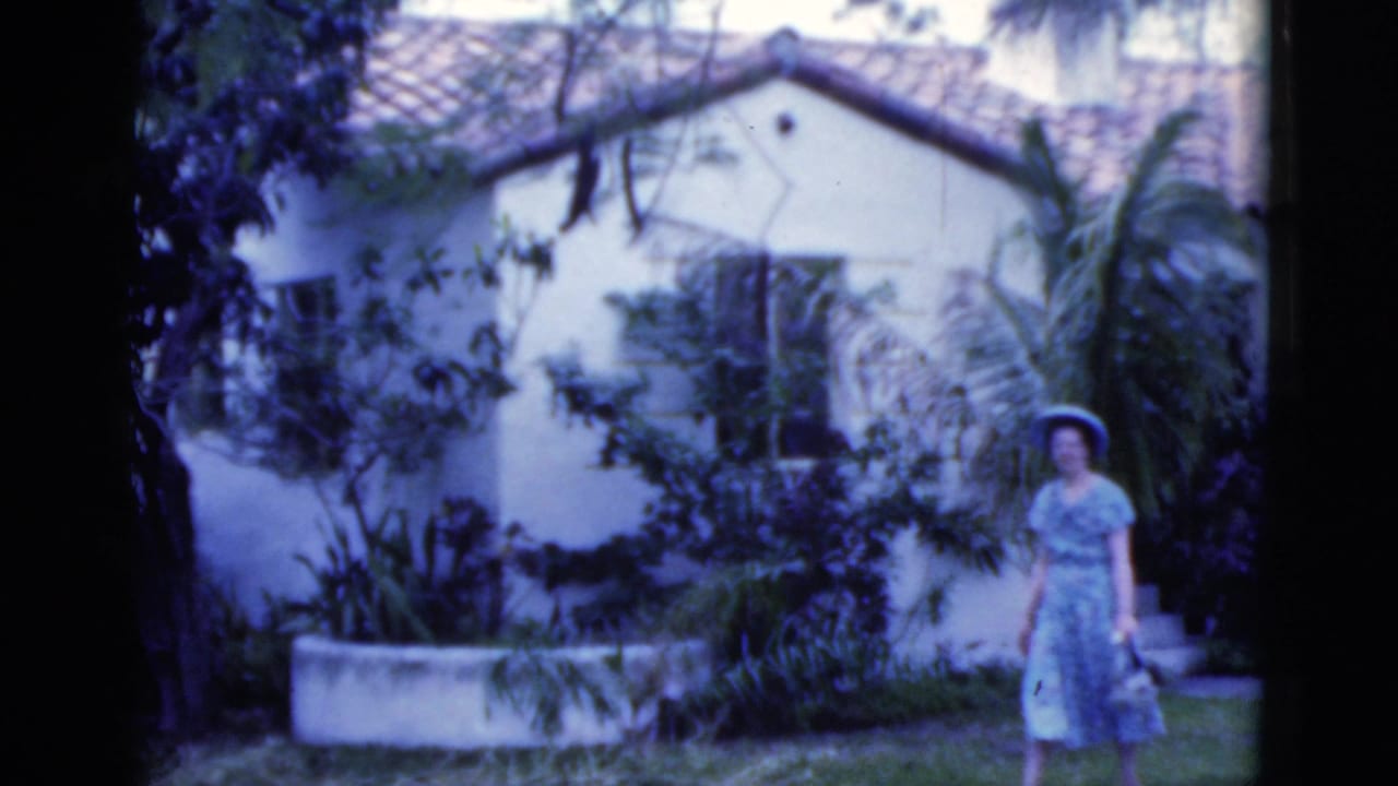 Woman in a blue dress and wide hat stands near a white stucco house behind garden foliage.