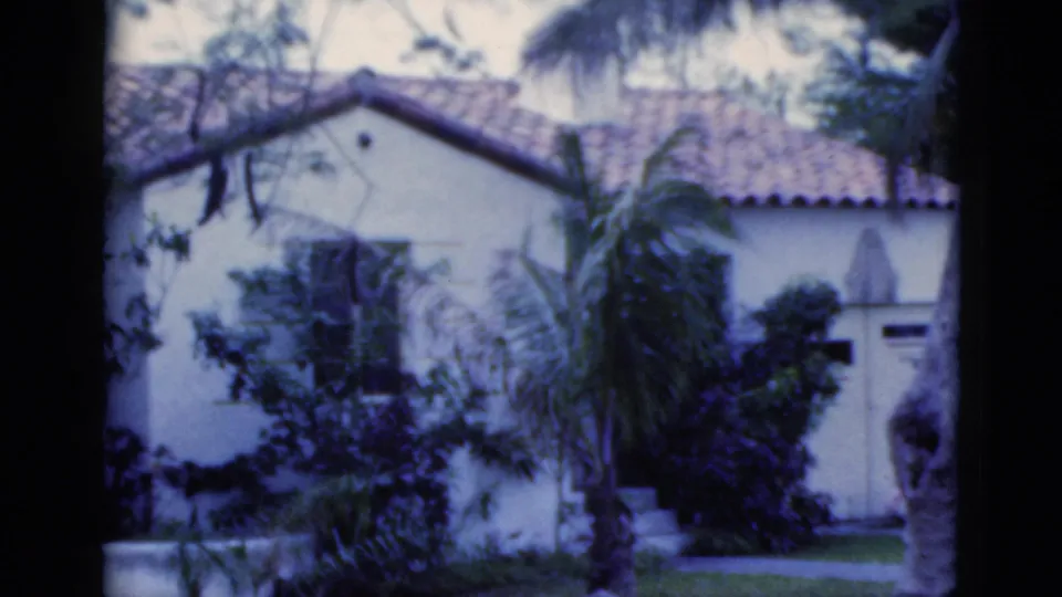 1975 Florida 8mm frame of a white stucco house with roof tiles, palms, shrubs, and a lawn seen from a yard.