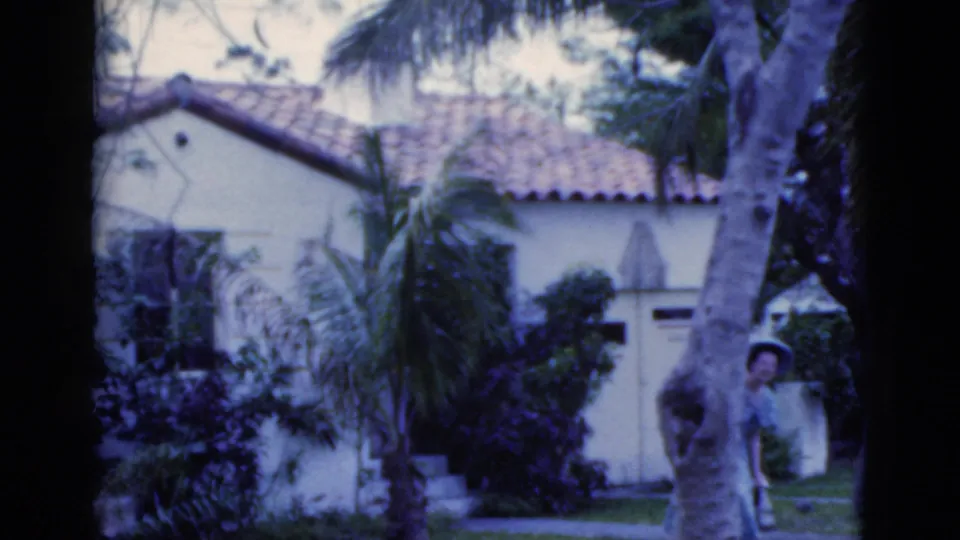 1975 Florida 8mm frame with a woman in a blue dress and wide hat peeking near a large tree beside a stucco house and palms.