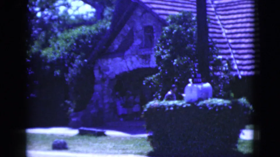 1975 Florida home-movie frame of the Parrot Jungle entrance area with a stone structure, hedges, lawn, and a small visitor figure.