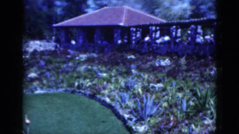 1975 Florida home-movie frame showing a red-roofed pavilion, stone terrace, visitors, tropical plantings, and a curved lawn edge.