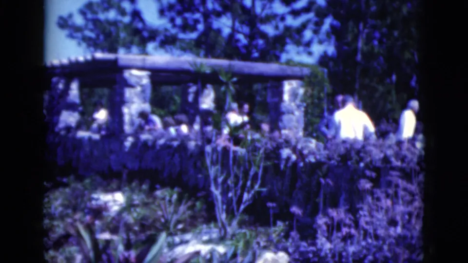 1975 Florida 8mm frame with visitors walking past a stone pergola in a lush outdoor resort-like garden.