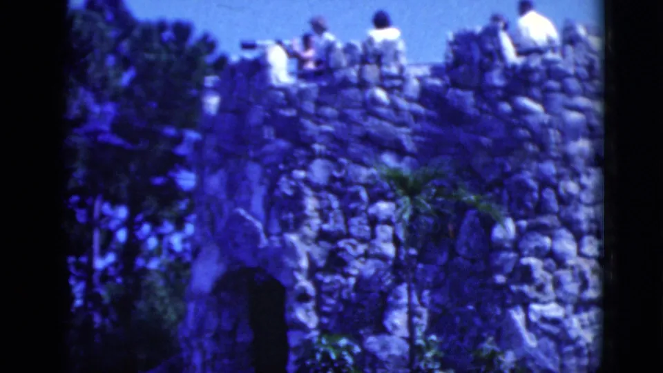 1975 Florida home-movie frame showing people gathered on a tall rock wall with stone texture and plants below.