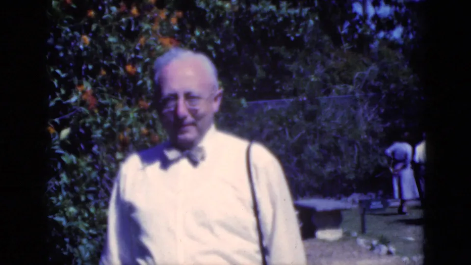 1975 Florida home-movie frame of an older man in a white shirt and bow tie facing the camera in a sunny garden.