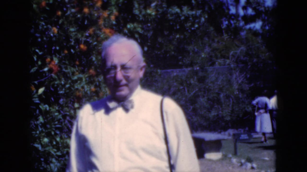 Older man in a light shirt and bow tie stands in a Florida garden, with orange fruit and distant walkers behind him.