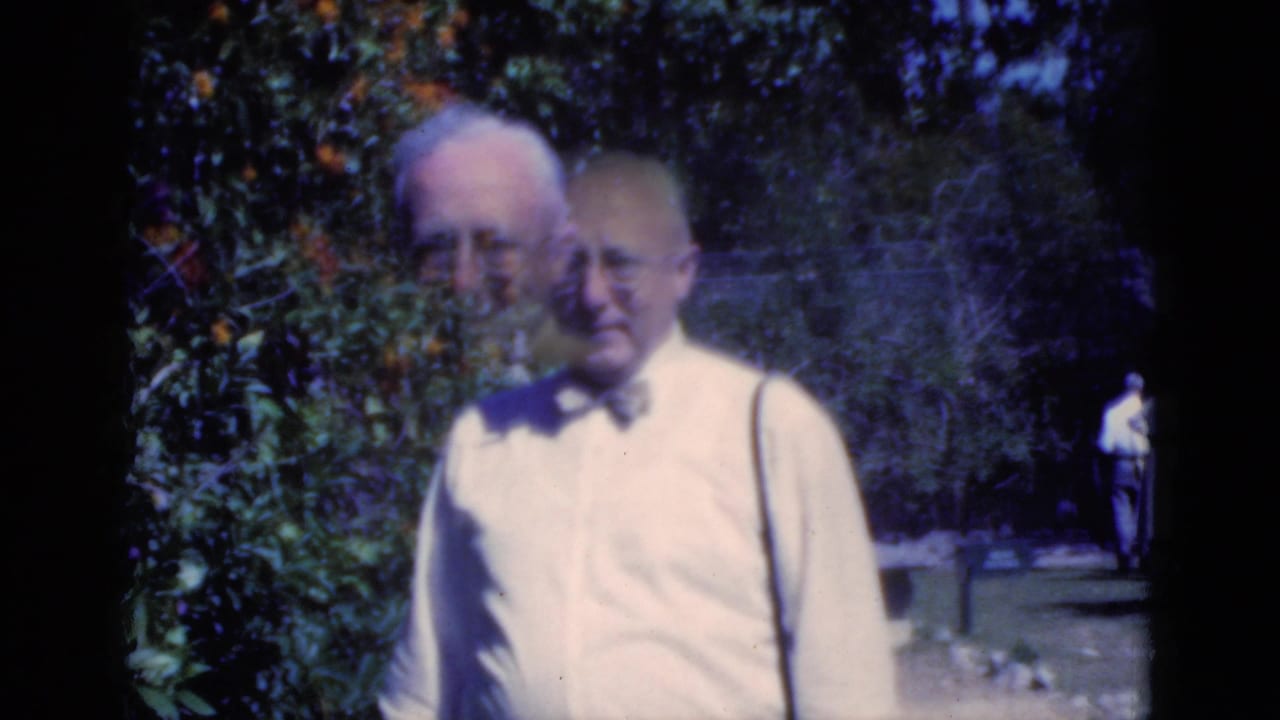 A 1975 Florida home-movie frame shows an older man in a bow tie near citrus branches, with a second face briefly overlapping in motion.