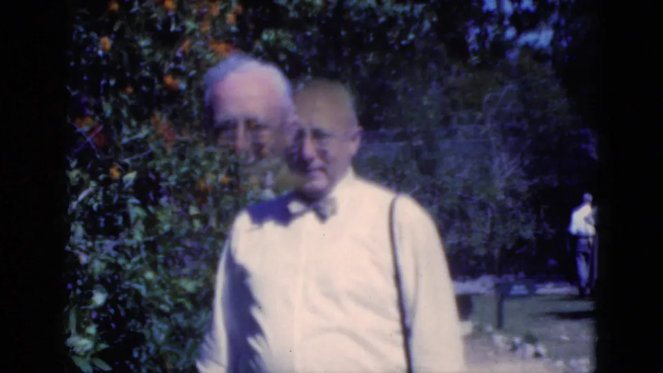A 1975 Florida home-movie frame shows an older man in a bow tie near citrus branches, with a second face briefly overlapping in motion.