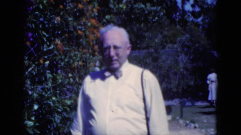 Older man in a light shirt and bow tie faces the camera in a Florida garden, with citrus foliage and two people in the distance.