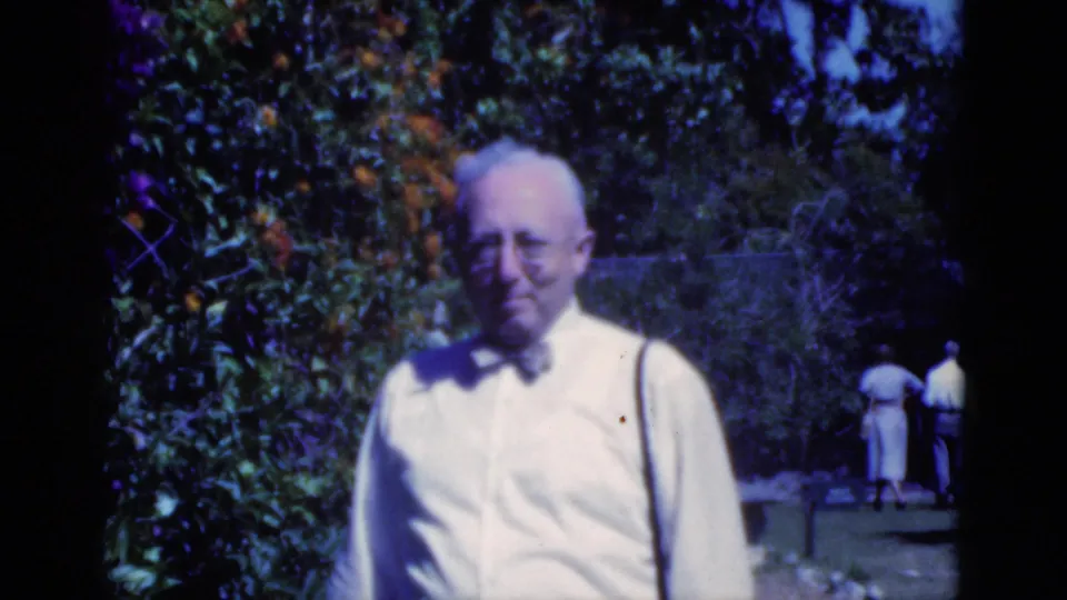 In this 1975 Florida garden frame, an older man stands in front of citrus foliage while two background figures walk away.