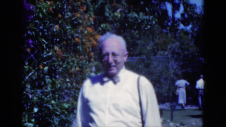 In 1975 Florida home-movie footage, a man in a white shirt, bow tie, and suspenders stands near dense garden foliage and orange blooms.